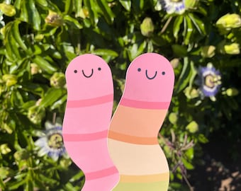 May include: Two colorful worm-shaped paper cutouts with happy faces. The worm on the left is pink with pink stripes. The worm on the right is rainbow colored with stripes of orange, yellow, green, blue, and purple.