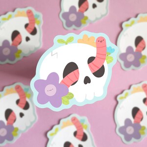 May include: A white skull sticker with a pink worm crawling out of the eye socket. The skull is surrounded by flowers and leaves in shades of purple, yellow, and green.