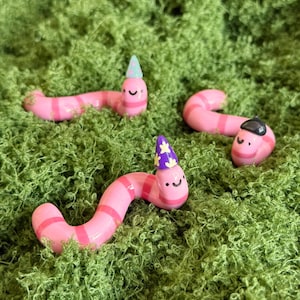 以下が含まれることがあります： 縞模様の体と笑顔の顔を持つ、3つの気まぐれなピンク色のミミズの置物。それぞれ小さな帽子をかぶっています。ミミズは鮮やかな緑色の苔を背景に配置され、遊び心のあるカラフルなシーンを作り出しています。