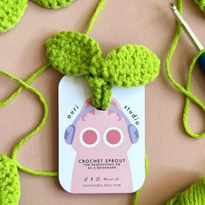 Op de afbeelding: Een handgemaakte groene gehaakte spruit bladwijzer of hoofdtelefoon accessoire. De spruit is bevestigd aan een label met een roze kat met een koptelefoon en de tekst "CROCHET SPROUT FOR HEADPHONES OR AS A BOOKMARK". De achtergrond is lichtroze.