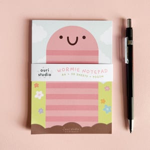 May include: Pink notepad with a cartoon worm design. The notepad has a striped pattern and says "Wormie Notepad A6 - 50 Sheets - 90GSM" on the front.