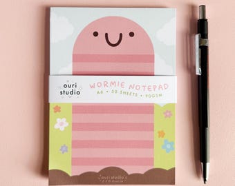 May include: Pink notepad with a cartoon worm design. The notepad has a striped pattern and says "Wormie Notepad A6 - 50 Sheets - 90GSM" on the front.