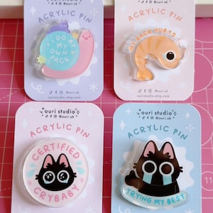 May include: Four acrylic pins on backing cards. The pins feature cartoon designs: a snail with text "I go at my own pace", a shrimp with text "My back hurts", a crying cat with text "Certified Crybaby", and a crying cat with text "Trying my best".