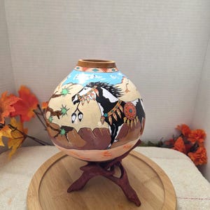 May include: A round, hollow gourd vessel featuring a hand-painted scene of a black and white horse in a desert landscape. The horse wears decorative gear with feathers. The gourd has a light blue sky, brown rock formations, and a wooden stand.