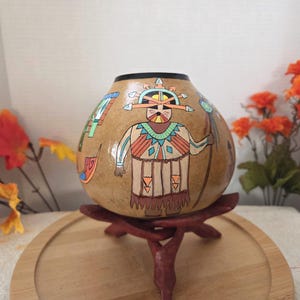 May include: A round, tan-colored gourd vessel featuring a hand-painted figure wearing a headdress and holding a staff. The figure is decorated with geometric patterns in shades of orange, teal, and black. The gourd has a dark rim.