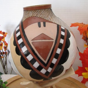 May include: A hand-painted gourd vase with a unique design. The vase features a tan base with a geometric pattern in shades of brown, black, and white. The top rim is brown, and the design includes a triangle shape. Decorative fall foliage surrounds the vase.