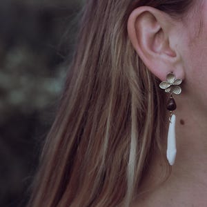 May include: A gold-coloured flower-shaped earring with a white, tooth-like pendant. The earring also features a dark brown, teardrop-shaped bead. The earring is worn on a person's ear, with long, light brown hair in the background.