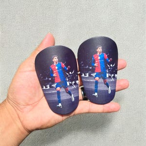 May include: Two black shin guards, each featuring a full-color image of a soccer player in a red and blue uniform. The player is running on a soccer field. The shin guards are held in a hand.