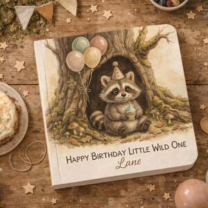 May include: A square book with a light-colored cover featuring an illustration of a raccoon wearing a party hat inside a tree hollow, holding a cupcake. Three balloons float above the raccoon. The text reads: Happy Birthday Little Wild One Lane.
