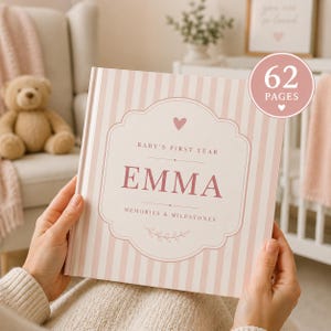 May include: A square baby memory book with a light pink and white vertical striped cover. The centre features a decorative frame with the text Baby's First Year, the name EMMA, and Memories & Milestones, topped with a small heart icon.
