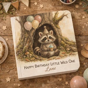 May include: A square book with a watercolor illustration of a raccoon wearing a party hat inside a tree hollow, holding a cupcake. Three balloons float above the raccoon. The text reads Happy Birthday Little Wild One and the name Lane.