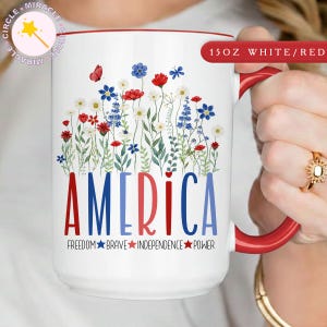 Op de afbeelding: Een witte keramische mok met een rode binnenkant en handvat, versierd met een bloemenontwerp en het woord "AMERICA" in rood en blauw. De mok bevat ook de woorden "FREEDOM BRAVE INDEPENDENCE POWER".