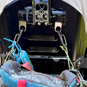 Può includere: Uno schienale per sedia a rotelle nero con una staffa di montaggio centrale in metallo dotata di leva. Due moschettoni neri sono fissati alla staffa, assicurando al telaio una borsa blu e grigia a griglia con cinghie rosse e cordini verde chiaro.