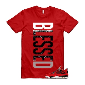 May include: A red t-shirt with the word "BLESSED" printed vertically in white, black, and gray. A red, black, and white sneaker is at the bottom of the shirt. The shirt is a crew neck style.