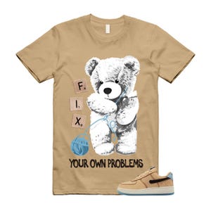 May include: Tan t-shirt featuring a white teddy bear holding a blue ball of yarn. The words "FIX" and "YOUR OWN PROBLEMS" are printed on the shirt. A pair of tan and blue sneakers are at the bottom.