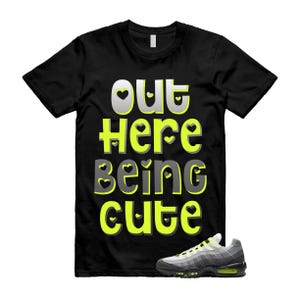 May include: Black t-shirt with the text "OUT Here Being Cute" in a playful font with heart accents. A pair of gray and black sneakers with neon green accents is placed at the bottom right.