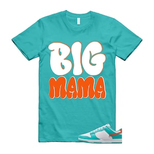 May include: A turquoise t-shirt with the words "BIG MAMA" in white and orange lettering. A pair of white, turquoise, and orange trainers is shown below the shirt. The shirt has a crew neck and short sleeves.