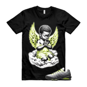 Pode incluir: Camiseta preta com um gr&aacute;fico de um anjo rezando com asas amarelas e um vestido amarelo, sentado em uma nuvem. Um par de t&ecirc;nis cinza e amarelo est&aacute; em primeiro plano.