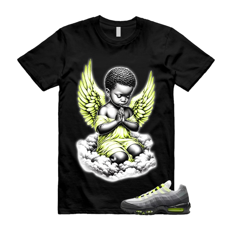 Pode incluir: Camiseta preta com um gr&aacute;fico de um anjo rezando com asas amarelas e um vestido amarelo, sentado em uma nuvem. Um par de t&ecirc;nis cinza e amarelo est&aacute; em primeiro plano.