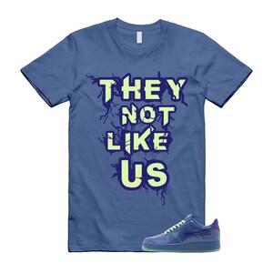 May include: A blue t-shirt with the text "THEY NOT LIKE US" in a light green font with a dark blue outline. A blue sneaker with a green sole is next to the shirt.