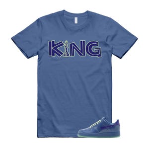 May include: Blue t-shirt with the word "KING" in a navy blue font with a chess piece graphic. A navy blue sneaker with a green sole is next to the shirt.
