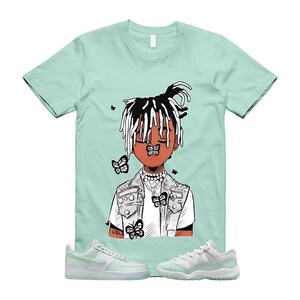 Puede incluir: Una camiseta verde claro con una ilustración de dibujos animados de una persona con mariposas. La persona tiene rastas blancas y negras, un chaleco blanco y un collar. Zapatillas deportivas verde claro a juego en la parte inferior. La camiseta tiene la palabra "WRL".