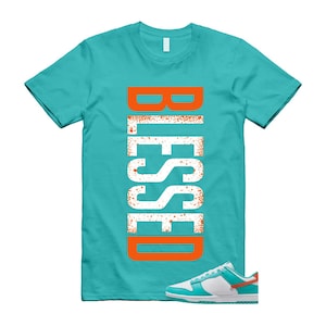 May include: A turquoise t-shirt with the word "BLESSED" in large, vertical, orange and white letters. A pair of white, turquoise, and orange trainers is at the bottom right of the image. The t-shirt is a crew neck.