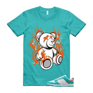 May include: A turquoise t-shirt with a white teddy bear graphic surrounded by orange flames and crossed-out eyes. The image also shows a pair of white, turquoise, and orange trainers.