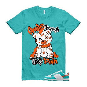 May include: A turquoise t-shirt featuring a white teddy bear graphic with orange detailing and the text "Smile Through The Pain." A pair of white, turquoise, and orange trainers are in the lower right corner.