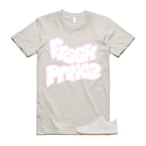 May include: A light beige t-shirt with the words "Fresh Prince" in white with a pink outline. A pair of light beige sneakers with a white sole and a gum-colored outsole are at the bottom of the image.