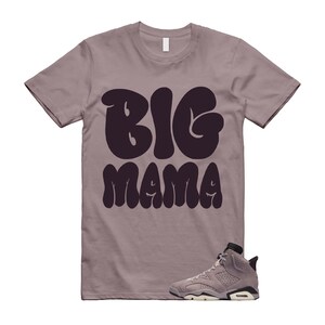 以下が含まれることがあります： 胸の中央に大きなダークパープルの丸みを帯びた文字で「BIG MAMA」とプリントされた、モーブカラーの半袖Tシャツ。右下隅には、Tシャツと同系色のモーブとダークパープルのハイカットスニーカーが配置されています。