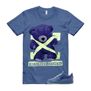 May include: Blue t-shirt with a purple teddy bear graphic wrapped in a neon green design. The text "THE STRUGGLE IS REAL" is below the bear. A blue sneaker with a purple swoosh is at the bottom.