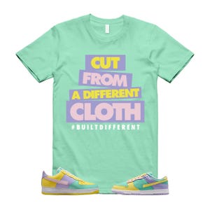 May include: A light green t-shirt with the text "CUT FROM A DIFFERENT CLOTH" in yellow, purple, and pink. Below the text is the hashtag "#BUILTDIFFERENT". The image also shows a pair of colorful sneakers.