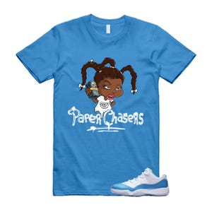 May include: A light blue t-shirt featuring a cartoon illustration of a girl holding money, with the text "Paper Chasers" in white. The image also shows a pair of white and blue sneakers.