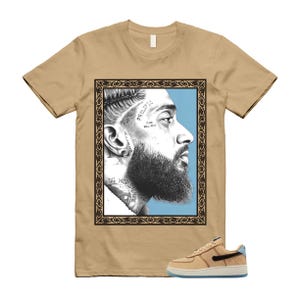May include: Tan t-shirt featuring a framed portrait of a man with a beard and tattoos. The portrait is set against a light blue background. A pair of tan and blue sneakers are in the foreground.