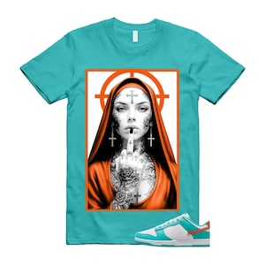 May include: A turquoise t-shirt with a graphic of a tattooed woman in an orange hood and crosses. The graphic is set against a white background with an orange target. White, turquoise, and orange trainers are in the image.