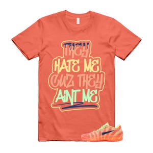 May include: A coral-colored short-sleeve t-shirt featuring the text "THEY HATE ME CUZ THEY AINT ME" in a multi-colored graffiti-style font. Below the text, a low-top sneaker with a coral and yellow patterned upper and a dark blue swoosh is visible.