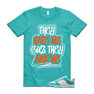 May include: Turquoise t-shirt with the text "THEY HATE ME CUZ THEY AINT ME" in white and orange. A pair of white, turquoise, and orange trainers are in the bottom right corner. The shirt is a crew neck style.