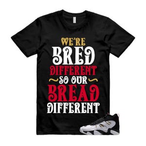 Può includere: Una t-shirt nera a maniche corte con il testo WE'RE BRED DIFFERENT SO OUR BREAD DIFFERENT in lettere bianche, rosse e dorate. Una scarpa sportiva nera e bianca con cinturino è posizionata in basso a destra sul design della maglietta.