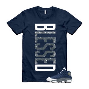 May include: A navy blue short-sleeve t-shirt featuring the word BLESSED printed vertically in large white letters with a distressed, speckled texture. A single navy blue and white athletic sneaker is positioned at the bottom right corner.
