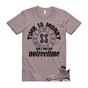 Puede incluir: Una camiseta malva con el texto "TIME IS MONEY" en un diseño de reloj, con símbolos de dólar en el centro y las palabras "and I ain't got nofreetime" debajo. Una zapatilla gris y blanca a juego está en la parte inferior.