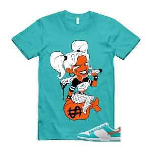 May include: A turquoise t-shirt with a cartoon illustration of a woman with white hair and orange skin, holding a bat and sitting on a money bag. The shirt is paired with white and turquoise trainers with orange accents.