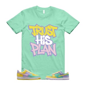 May include: A mint green t-shirt with the text "TRUST HIS PLAN" in yellow, white, and purple. Below the shirt are two pairs of colorful sneakers with yellow, purple, and blue accents.