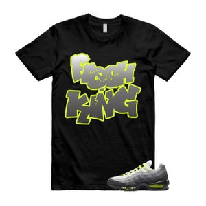 Op de afbeelding: Zwart T-shirt met de tekst "Fresh King" in een graffiti-stijl, omlijnd in neon groen. Een paar grijze, zwarte en neon groene sneakers staan onderaan de afbeelding.