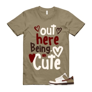 May include: A tan short-sleeve t-shirt featuring the text "out here Being Cute" in a mix of white and dark red lettering, accented with heart illustrations in red, white, and dark brown, paired with a white and tan low-top sneaker.