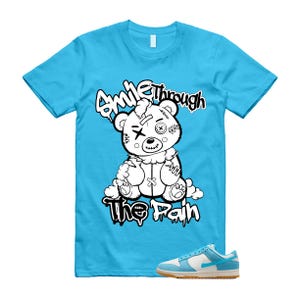 Könnte beinhalten: Hellblaues T-Shirt mit einer Grafik eines vernähten Teddybären und dem weißen Schriftzug "Smile Through The Pain". Ein Paar blau-weiße Sneaker mit Gummisohle befindet sich in der unteren rechten Ecke.