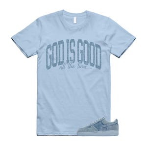 May include: Light blue t-shirt with the text "GOD IS GOOD all the time" in a textured, arched font. A pair of light blue sneakers with a matching textured design are at the bottom of the image. The t-shirt and sneakers are on a white background.
