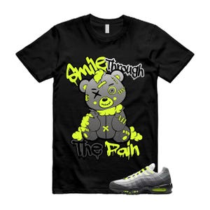 以下が含まれることがあります： 黒いTシャツ。 灰色のテディベアのグラフィックとネオンの緑色のアクセント、そして「Smile Through The Pain」というテキスト。 グレーとネオンのグリーンスニーカーも画像に表示されています。