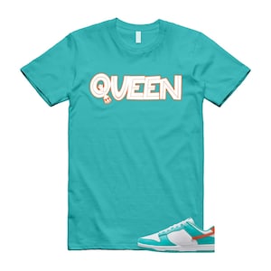May include: A turquoise t-shirt featuring the word "QUEEN" in white with orange outline. A matching turquoise, white, and orange trainer is positioned below the shirt. The shirt and trainer are set against a white background.