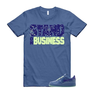 May include: Blue t-shirt with the text "STAND ON BUSINESS" in a distressed font. A blue sneaker with a purple swoosh is displayed next to the shirt. The sneaker has a green sole.